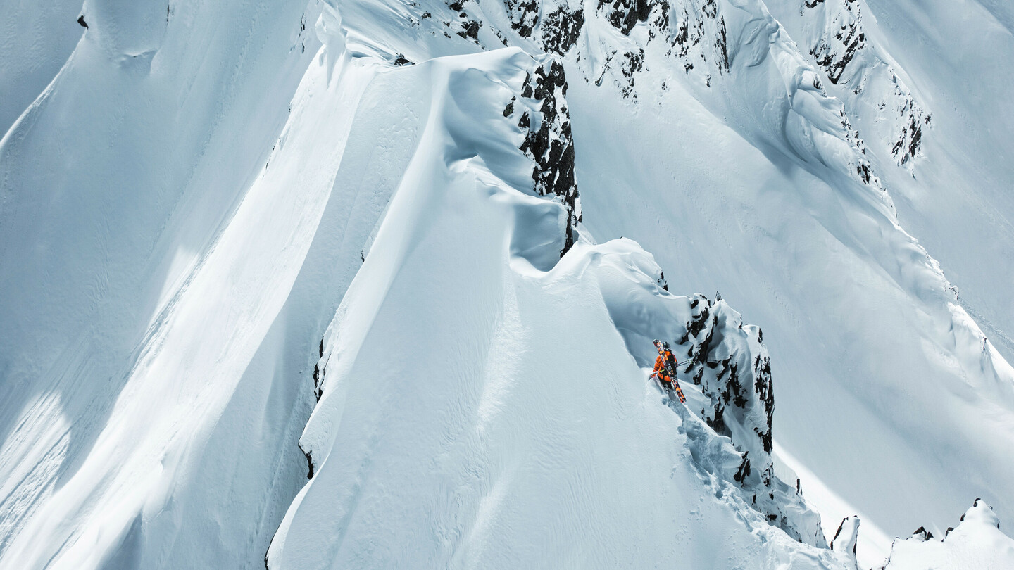 Skier in an orange jacket carefully navigating along a narrow, steep snow ridge in a dramatic mountain landscape.