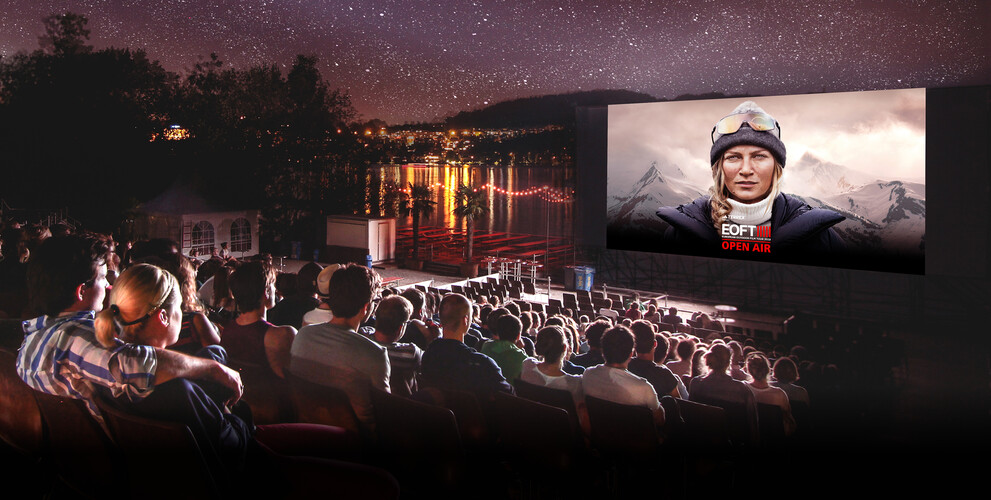 An outdoor cinema by the lake with a large audience watching a film under the starry sky. On the screen is a woman in outdoor gear with mountains in the background, and the text “EOFT OPEN AIR” is displayed.